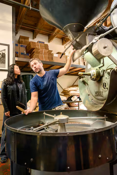Two people working with a large coffee roasting machine in a warehouse setting.