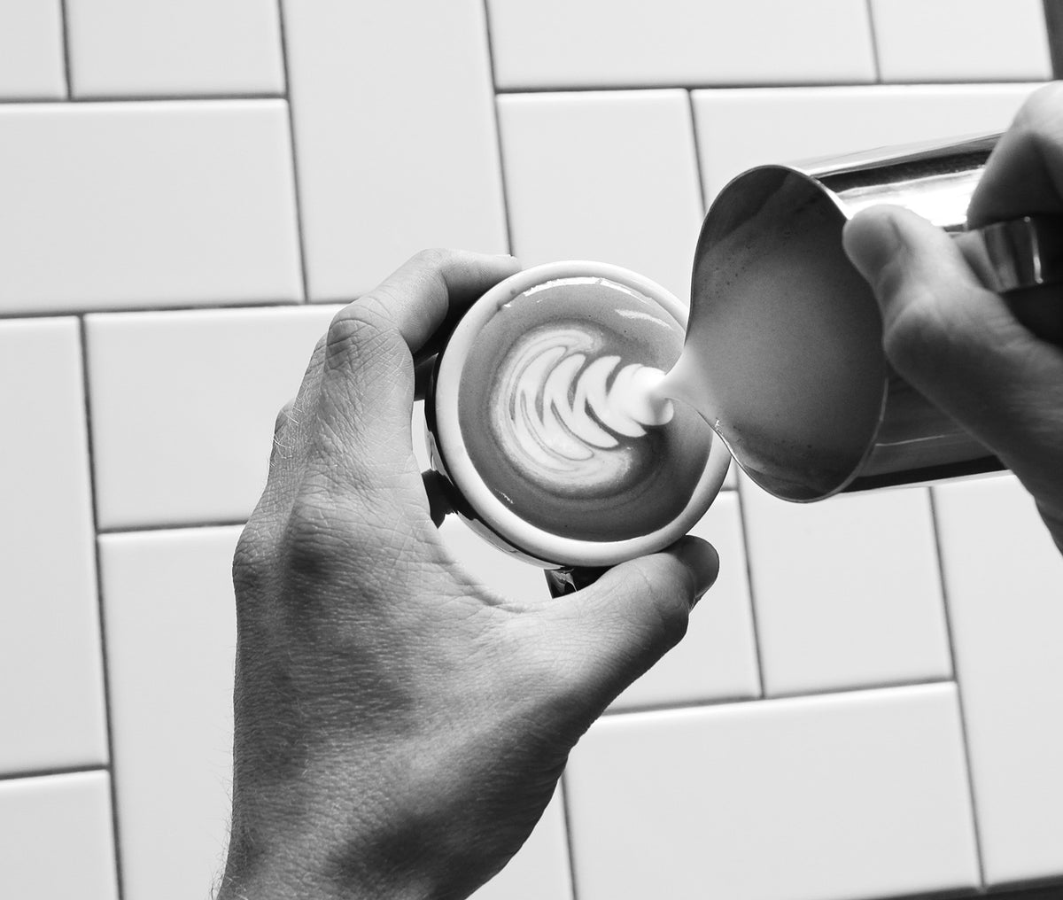 Hands pouring steamed milk from a metal pitcher into a cup of coffee to create latte art.