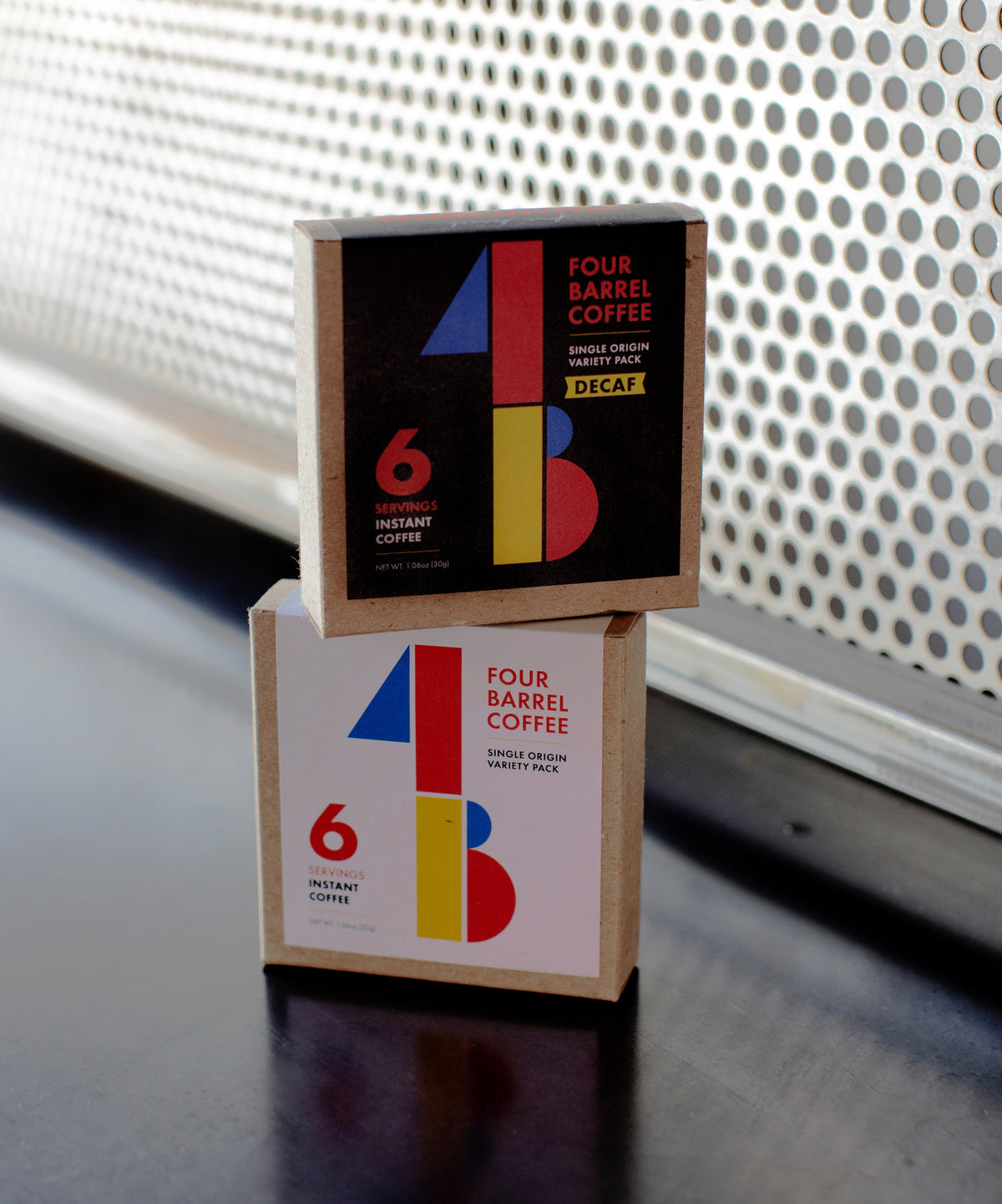 Two square boxes of INSTANT COFFEE with colorful geometric designs stacked on a dark surface.