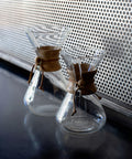Two clear glass coffee makers with brown leather straps on a reflective surface.