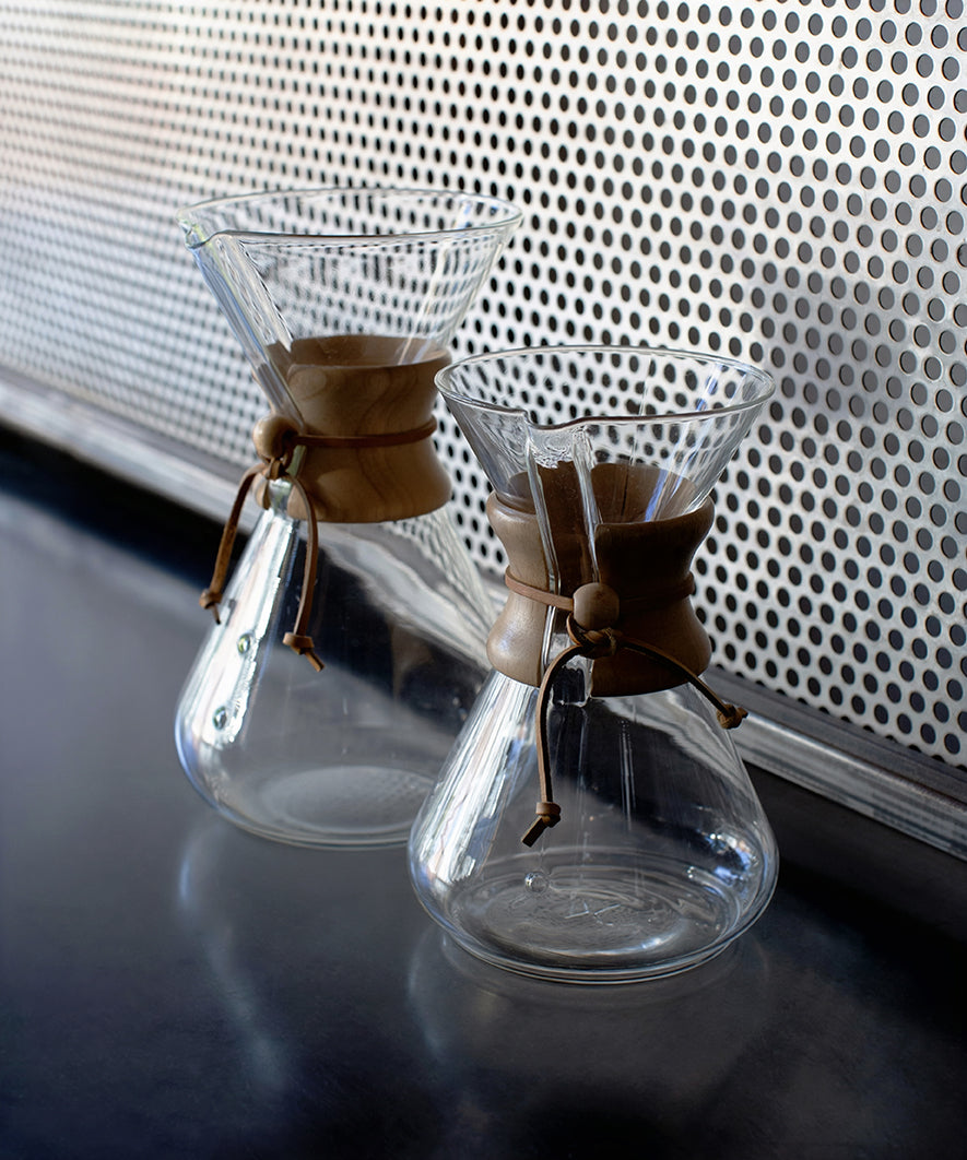 Two clear glass coffee makers with brown leather straps on a reflective surface.