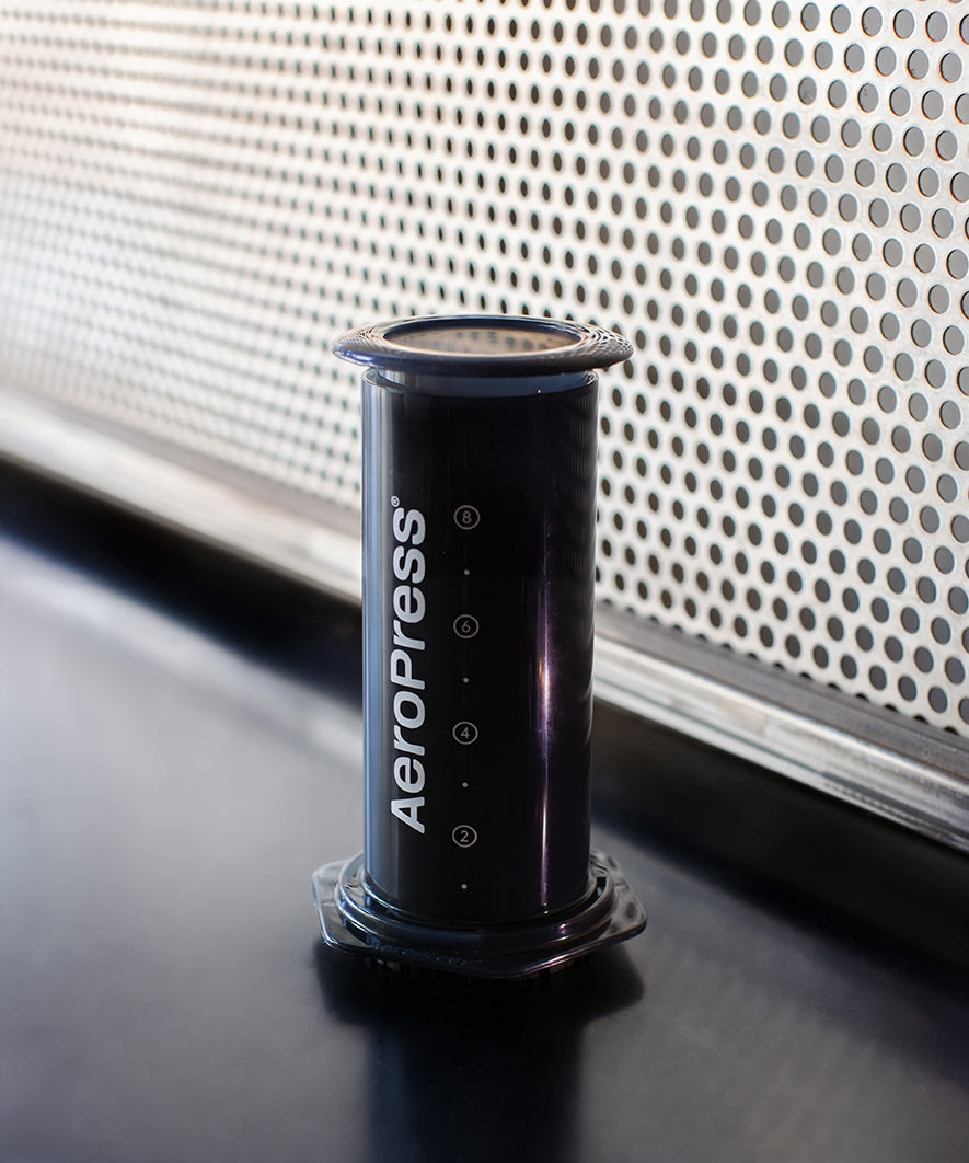 Black Aeropress coffee maker on a reflective surface with a perforated wall in the background