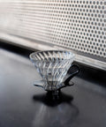 Clear glass coffee filter with black plastic handle on a dark surface.