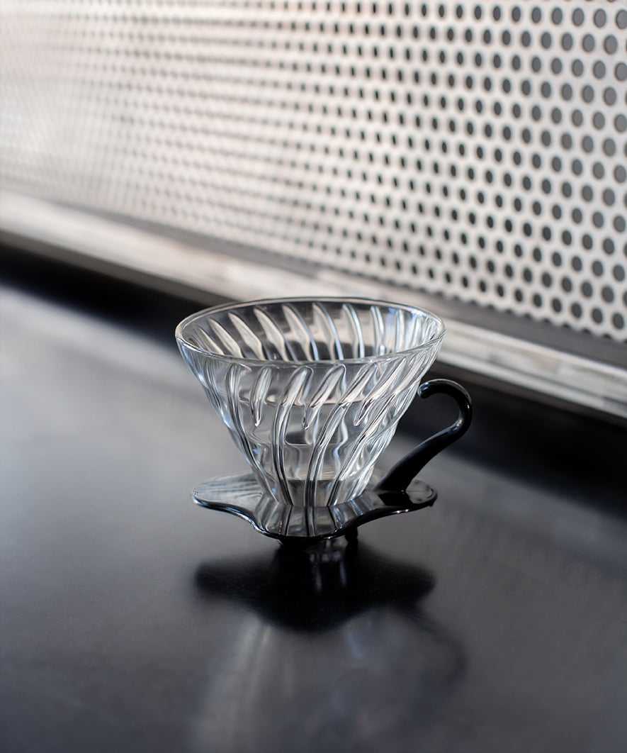 Clear glass coffee filter with black plastic handle on a dark surface.