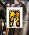 A rectangular coffee label or packaging card for 'four barrel coffee' with stylized artwork by Nat Russell, placed on a black mesh surface. The card has a white border and features a black central panel with yellow and orange starburst shapes and a white cat silhouette.