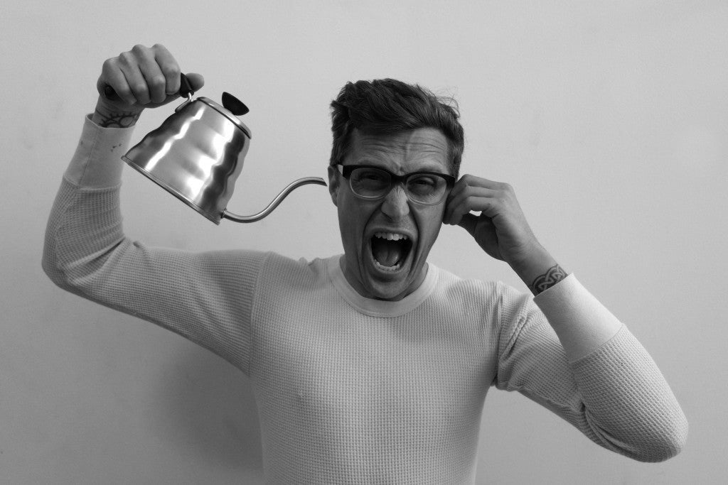 A man in glasses with his mouth open in an intense expression, holding a Hario Buono Kettle above his head.