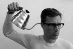 A man in glasses holds a stainless steel Hario Buono Kettle near his ear.