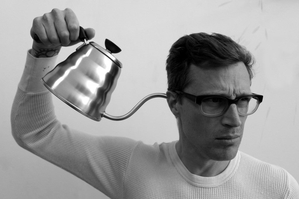 A man in glasses holds a stainless steel Hario Buono Kettle near his ear.