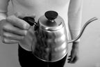 A person holds a stainless steel Hario Buono Kettle by its handle.