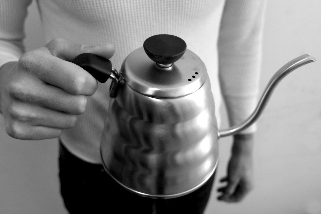 A person holds a stainless steel Hario Buono Kettle by its handle.