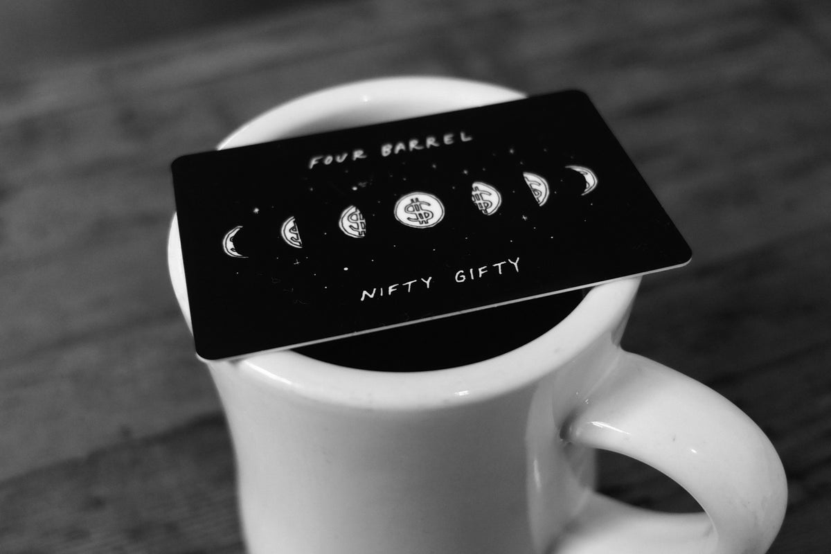 A black physical gift card featuring a lunar cycle of dollar signs resting across the rim of a white coffee mug.