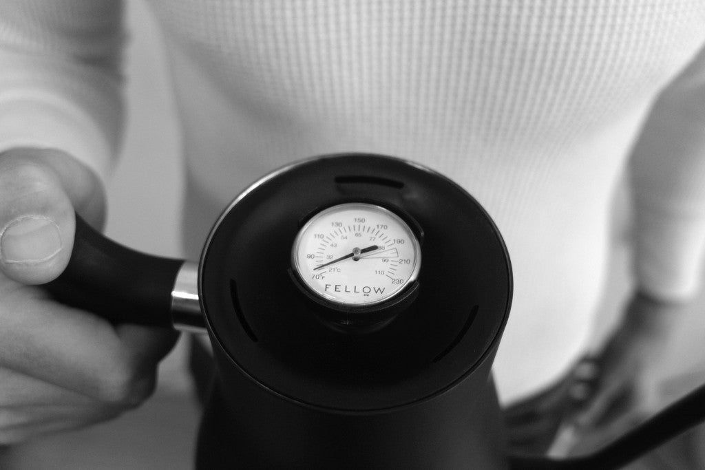 Close-up of a person holding a black Fellow Stagg Stove Top Pour Over Kettle with a built-in circular analog thermometer on its lid.