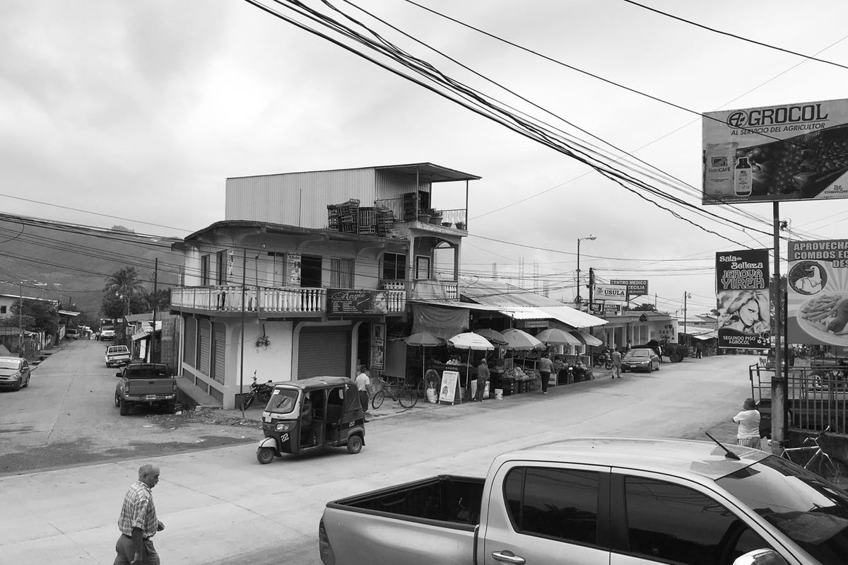A street scene in a rural town featuring a multi-story building, a parked auto rickshaw, small shops with signage, and a bag of HONDURAS ROBERT FIGUEROA ESPRESSO along a paved road.
