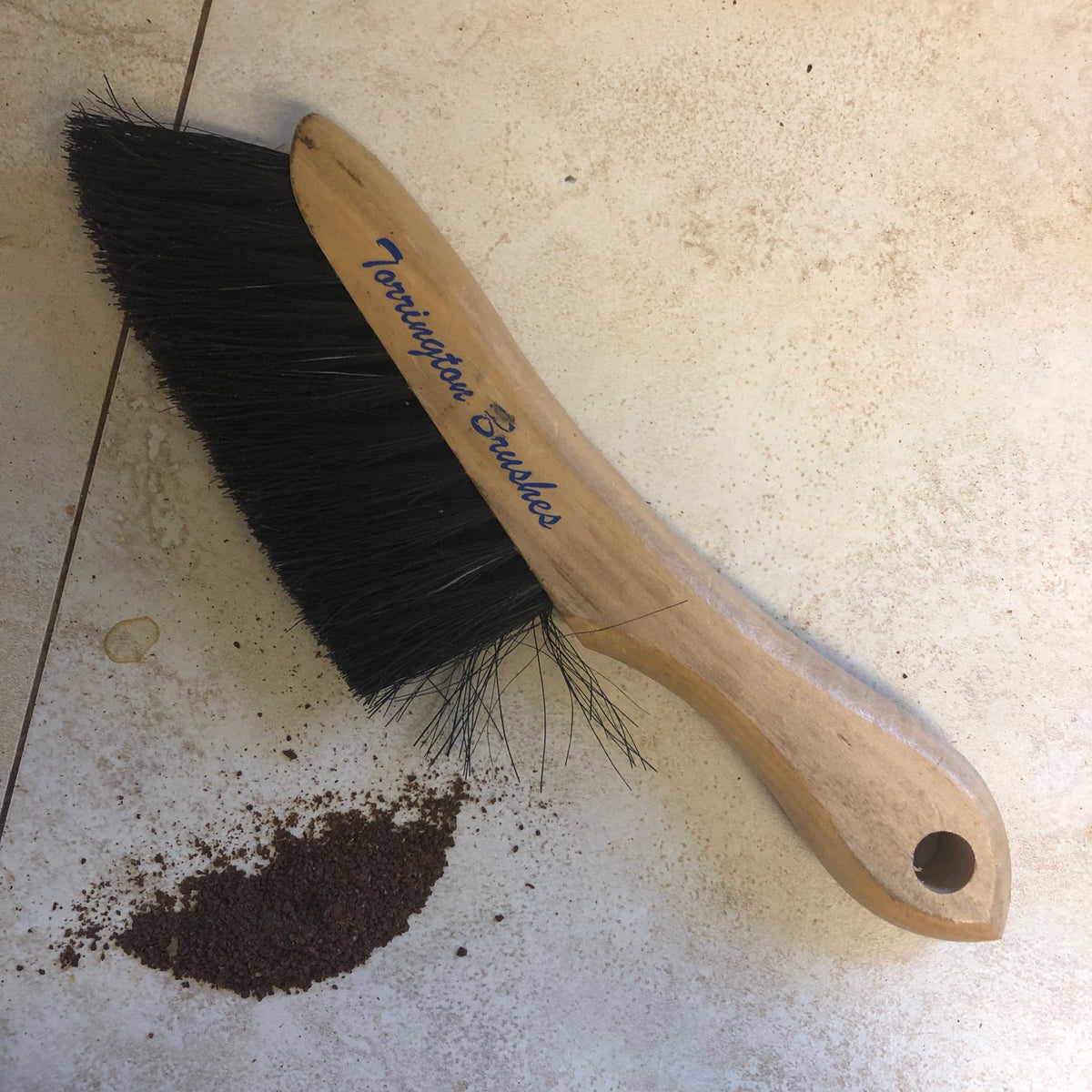 A torrington wood counter brush with black bristles lying on a tiled surface next to a small pile of dark debris.